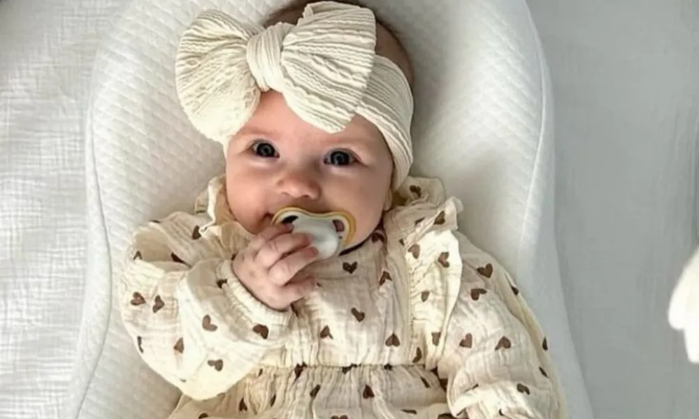baby shower trivia questions featured in a photo of a cute infant wearing a cream bow headband and a heart-patterned outfit holding a pacifier.