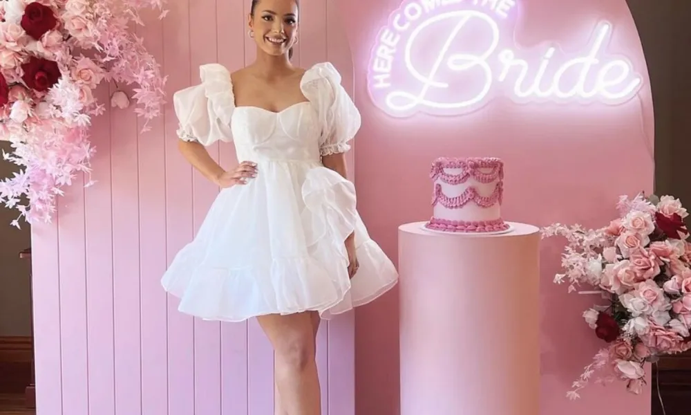 bridal shower trivia questions featured in a photo of a bride-to-be posing in a white puff-sleeve mini dress before a pink arch backdrop with a neon sign and floral arrangements.