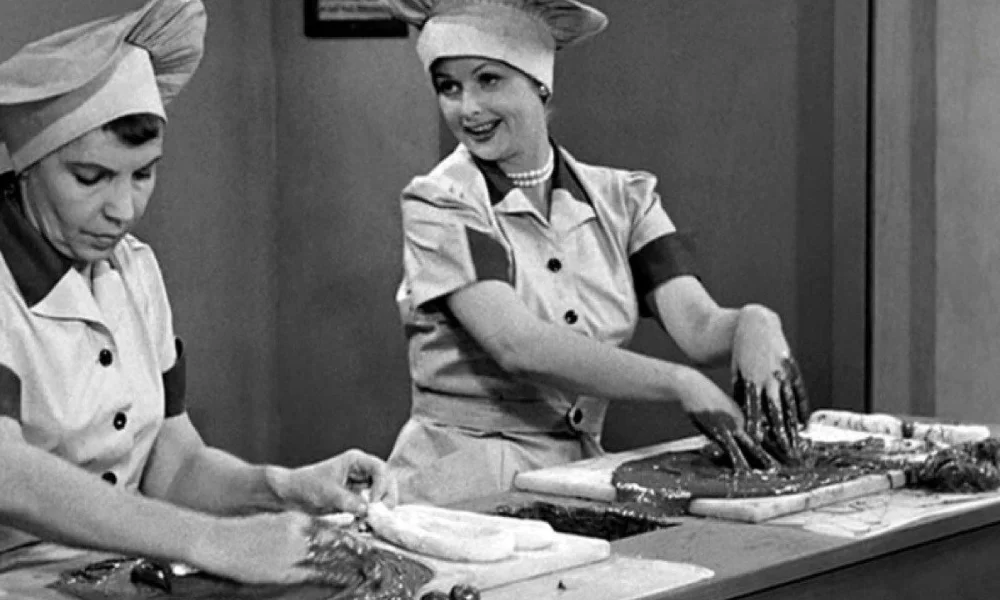 A vintage black-and-white image of Lucille Ball and Vivian Vance as Lucy and Ethel from I Love Lucy, wearing chef hats and uniforms while working at a candy factory table, perfect for testing your knowledge on classic tv trivia questions. 